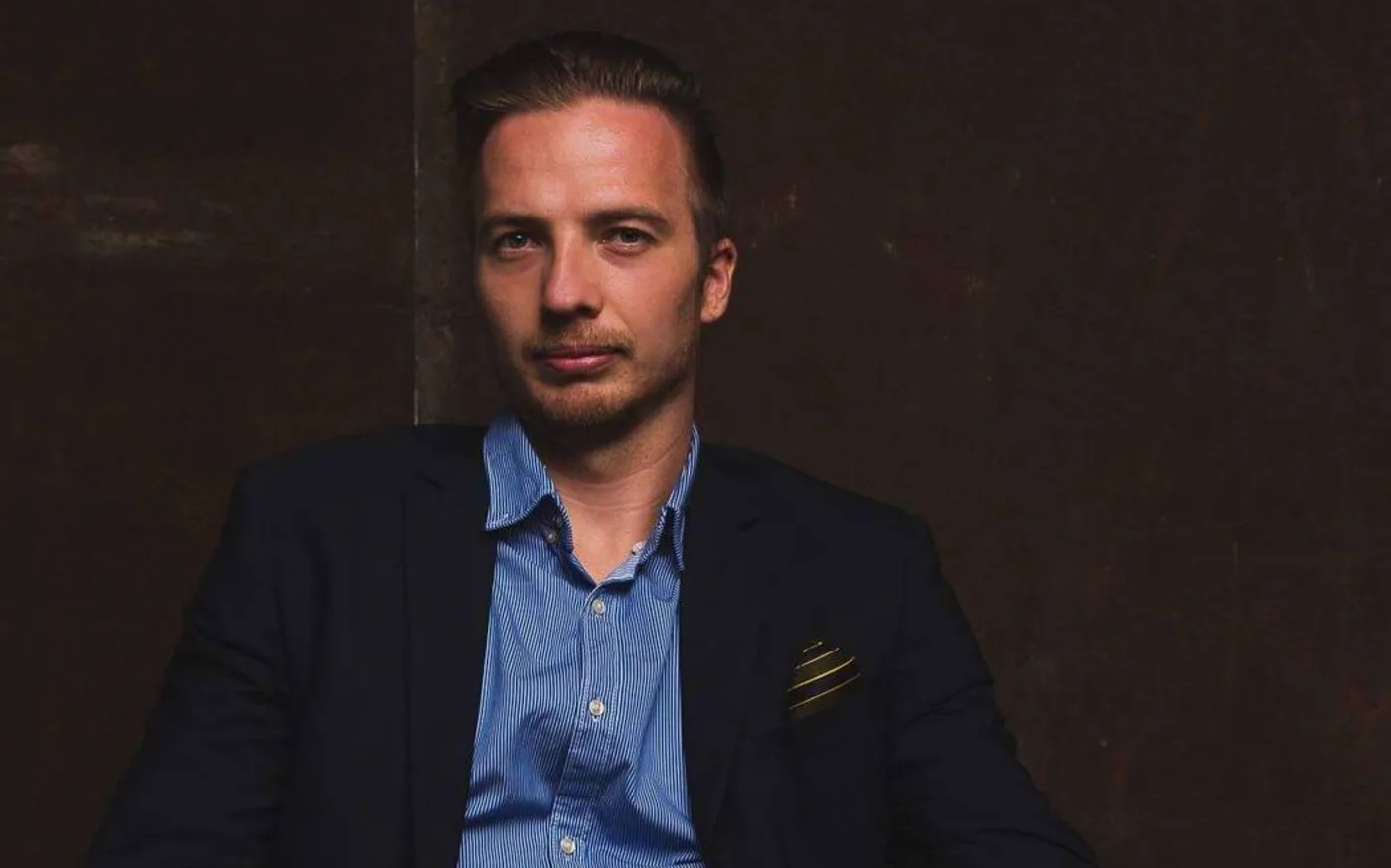 Ortox in a dark blazer and open blue shirt against a dark wood wall