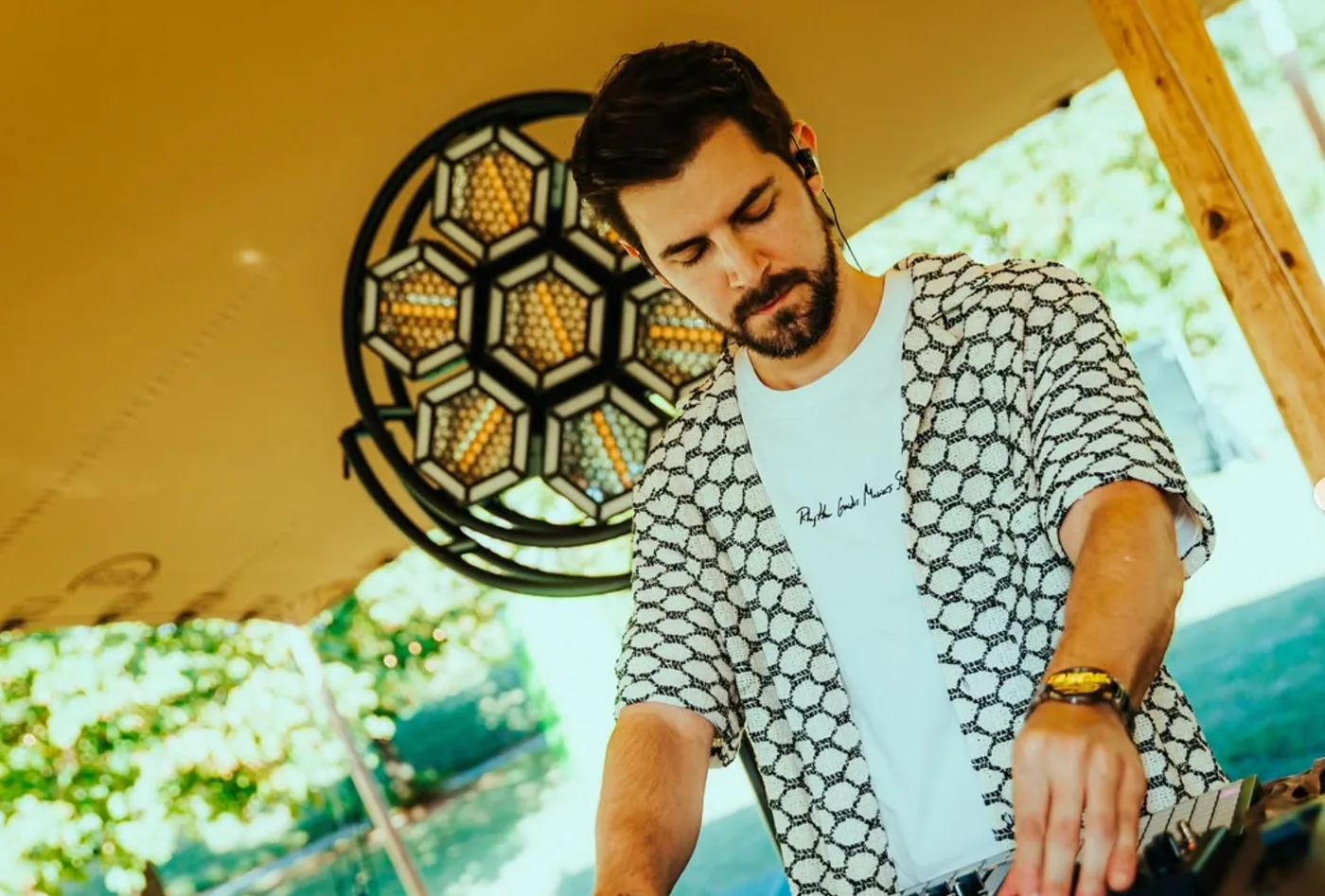 RTIK DJing outdoors under a wooden canopy in a patterned shirt