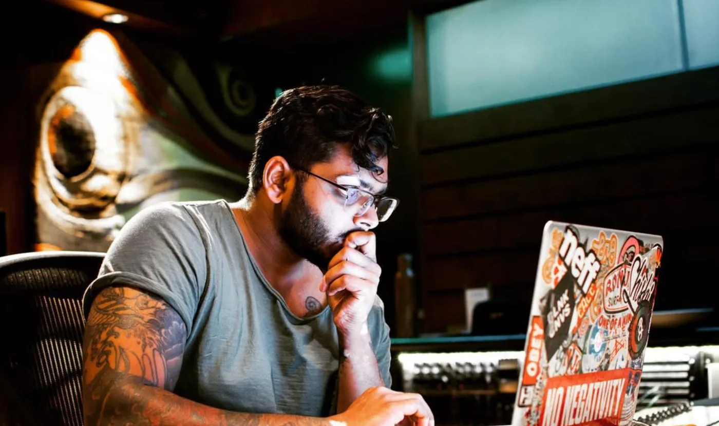 VNDL in his studio, chin in hand, looking at a sticker-covered laptop