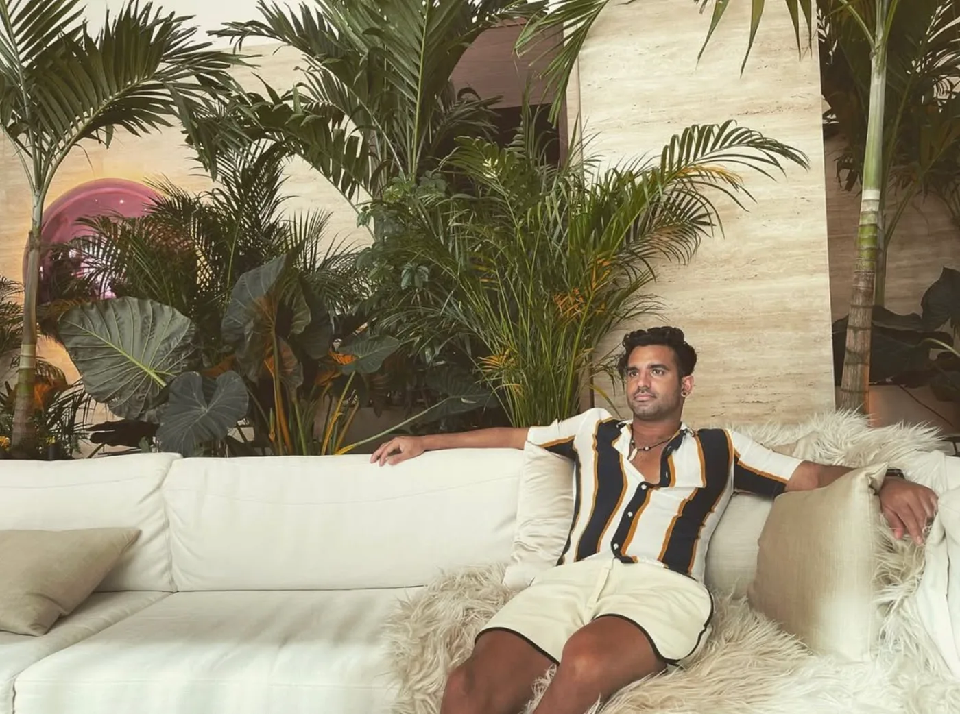 Flowaver in a striped polo lounging on a white couch surrounded by tropical plants and travertine