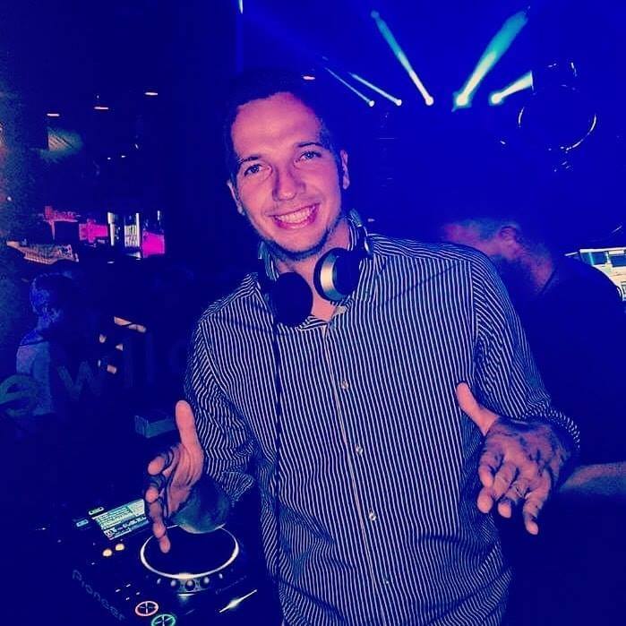 Ortox behind the decks in a club, headphones around his neck, hands raised mid-set