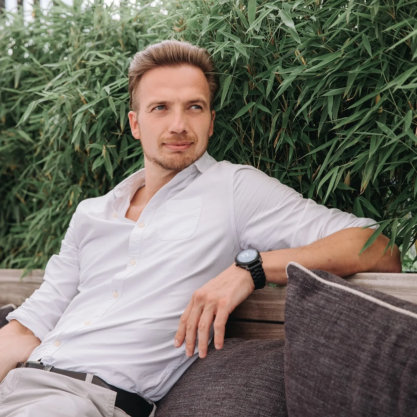 Ortox in a white shirt leaning back on an outdoor bench surrounded by bamboo
