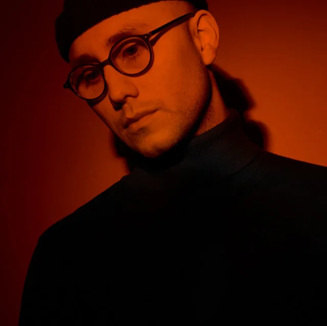 Sam Welch in a moody red-lit press portrait, beanie and glasses