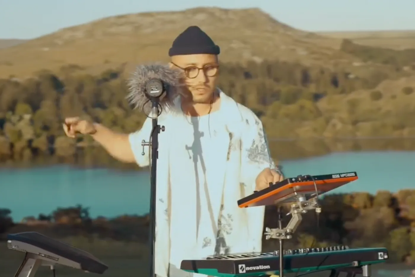 Sam Welch performing live outdoors by a lake, mic and keys in hand