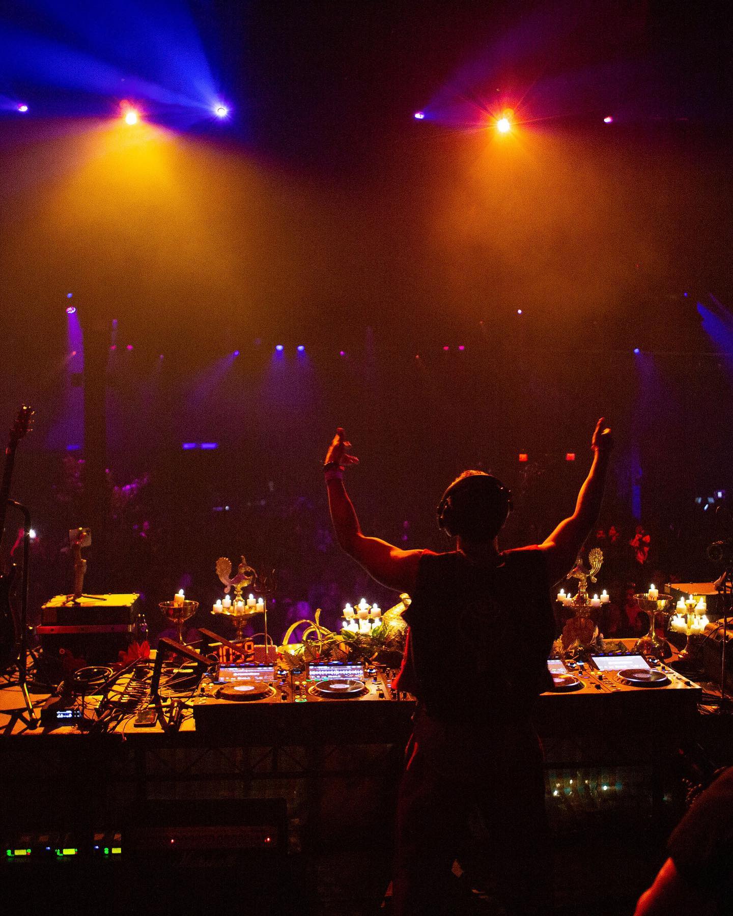 Shishi with arms raised behind the decks, stage lights blazing over a packed crowd