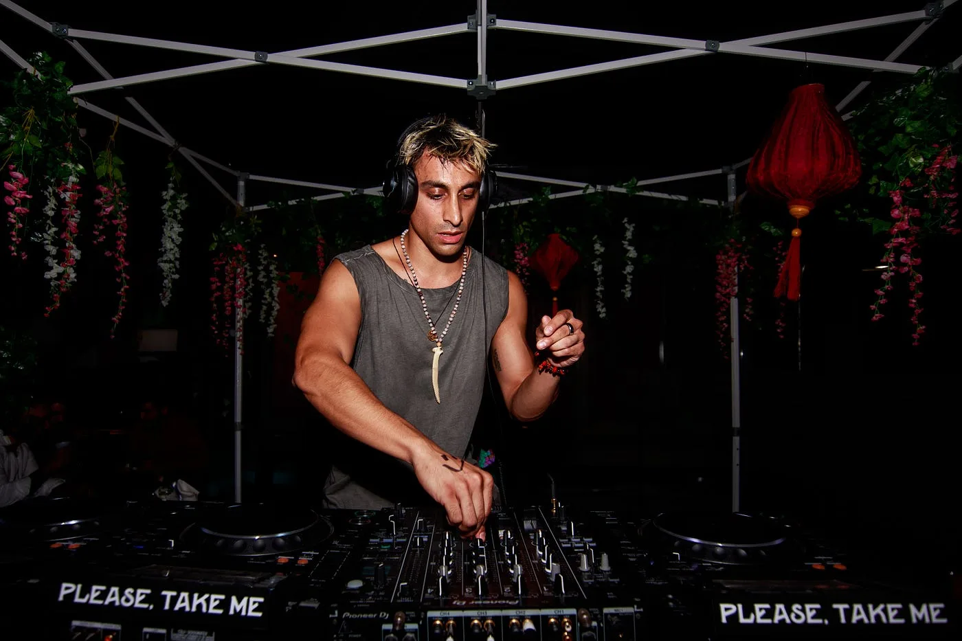 Shishi behind the decks in a tank top, headphones on, surrounded by hanging lanterns and flowers