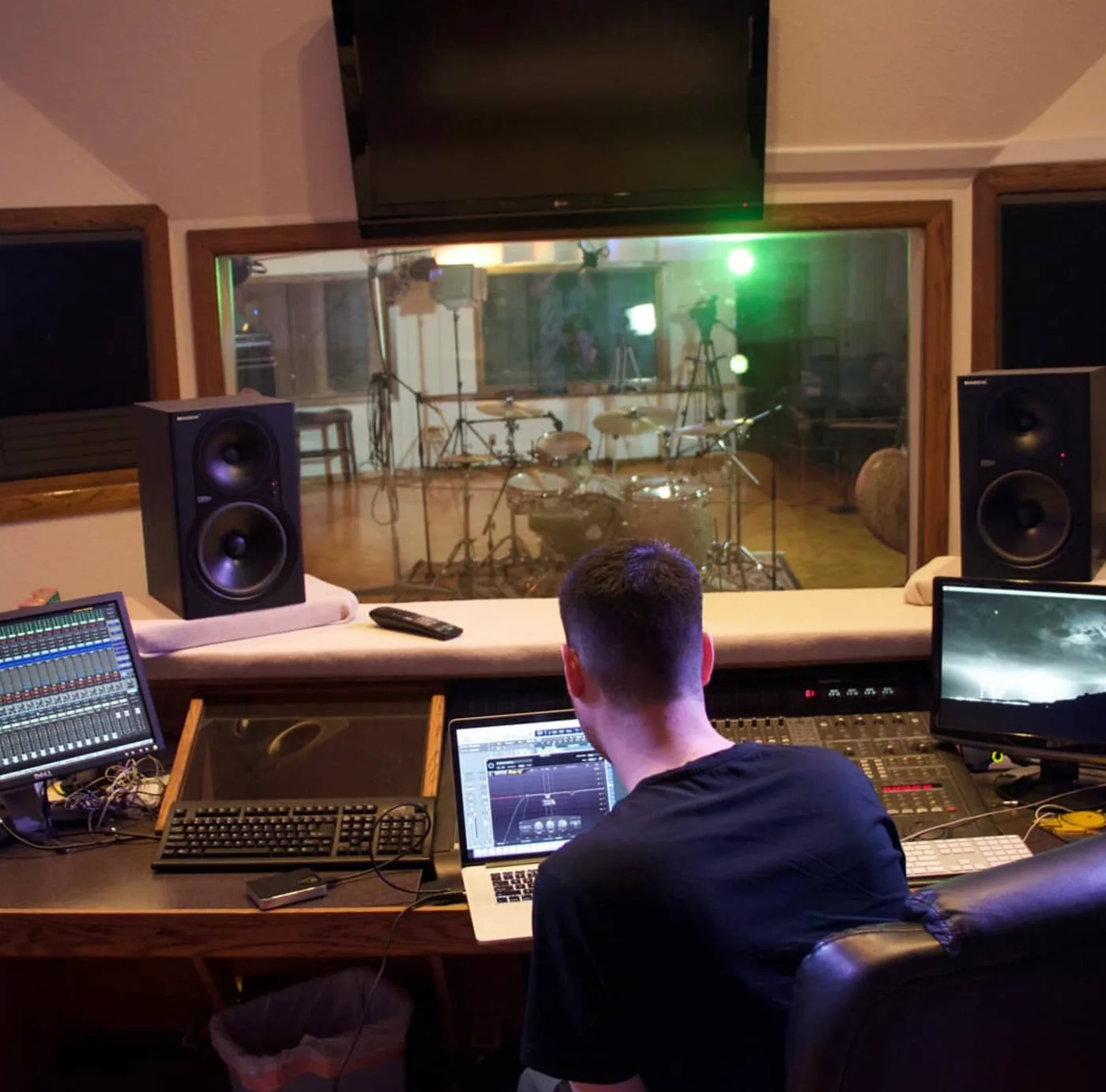 Tom Rogers working at a mixing desk in a pro studio control room with a live drum room behind him