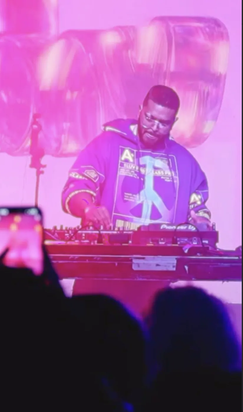 VNDL mixing live behind Pioneer CDJs in a hooded jacket, a fan filming on a phone in the foreground